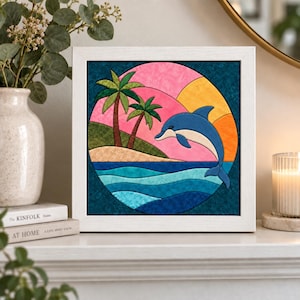 Op de afbeelding: Een vierkante witte lijst met een cirkelvormig stoffen kunstwerk dat een blauwe dolfijn toont die uit de golven springt. De scène bevat een zanderig eiland met twee palmbomen, een roze lucht en een gele zon tegen een donkerblauwe achtergrond.