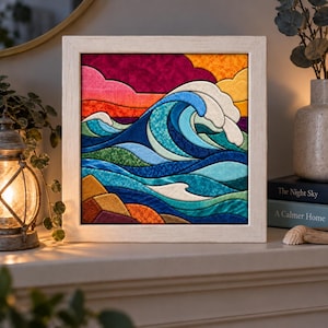 May include: A square framed fabric art piece featuring a stylized ocean wave in shades of blue and teal. The composition includes a vibrant sunset sky with clouds in red, orange, and yellow, and textured fabric sections forming the sea and waves.