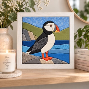 Könnte beinhalten: Ein gerahmtes Stoffkunstwerk mit einem Papageientaucher auf einem felsigen Ufer. Der Vogel hat einen schwarz-weißen Körper und einen orangefarbenen Schnabel. Der Hintergrund zeigt grüne Hügel, ein blaues Meer und einen hellblauen Himmel. Weißer Holzrahmen.