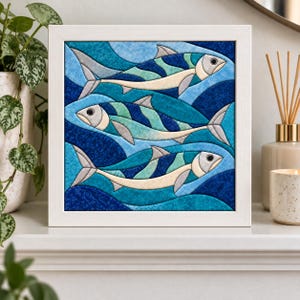 Op de afbeelding: Een vierkant ingelijst kunstwerk aan de muur met een stoffen quiltontwerp van drie vissen die in een blauwe oceaan zwemmen. De vissen zijn uitgevoerd in tinten crème, lichtblauw en donkerblauw tegen een achtergrond van golvende blauwe golven.