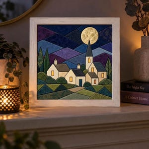 Op de afbeelding: Een vierkant ingelijst wandkunstwerk met een gestileerd nachtlandschap met een volle maan, een kerktoren en verschillende kleine huizen. De scène gebruikt geometrische vormen in tinten blauw, paars en groen, met warm geel licht uit de ramen.