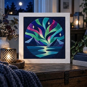 Puede incluir: Una pieza de arte mural cuadrada enmarcada que presenta un diseño circular de aplique de tela de la aurora boreal en tonos verdes, morados y azules sobre siluetas de montañas oscuras y un cuerpo de agua reflectante, con tres estrellas blancas.