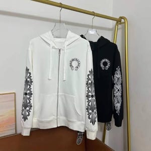 May include: A white zip-up hoodie and a black zip-up hoodie, both featuring a circular Chrome Hearts logo on the left chest and intricate black or white floral scrollwork patterns running down the length of each sleeve with a front pocket.