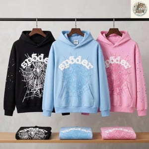 May include: Three hooded sweatshirts in black, light blue, and pink, each featuring a white spiderweb graphic on the front and back with the text sp5der. The hoodies are displayed hanging on hangers and folded on a shelf below.