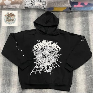 Könnte beinhalten: Ein schwarzer Kapuzenpullover mit einer großen weißen Grafik auf der Brust, die ein Spinnennetz mit dem darüber gewölbten Schriftzug "sp5der" zeigt. Die Ärmel sind mit kleinen weißen Sternen verziert und das Kleidungsstück hat eine Kapuze.