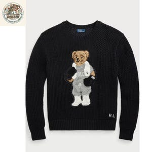 Può includere: Un maglione nero a maniche lunghe con girocollo e motivo a maglia testurizzato. Al centro, un orso ricamato indossa una felpa grigia con la parola POLO, pantaloncini grigi, stivali bianchi e guanti da boxe neri. Le lettere RL si trovano in basso.