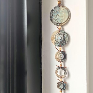 May include: A vertical wall hanging featuring six circular ceramic discs of varying sizes, connected by brown leather cords and accented with small wooden beads. The discs display blue, brown, and cream glazes with embossed patterns.