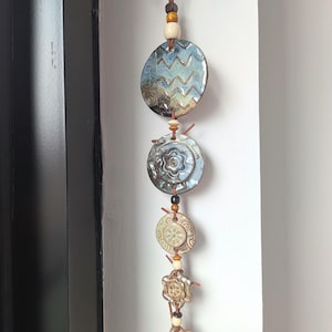 May include: A vertical wall hanging featuring five circular ceramic discs in varying sizes, suspended on a brown leather cord with wooden beads. The discs display blue, tan, and brown glazes with embossed patterns including zigzags and flowers.