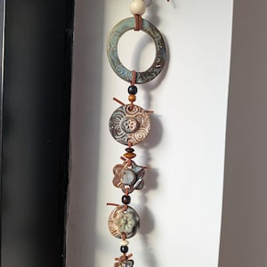 May include: A vertical wall hanging featuring a series of ceramic pieces in shades of blue, brown, and cream. The elements include a large ring, circular discs, and flower-shaped charms, all strung together with brown leather cord and small wooden beads.