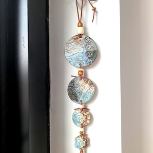 May include: A vertical wall hanging featuring five graduated ceramic discs in shades of blue, brown, and cream. The discs are strung together with brown leather cord and interspersed with small wooden beads, ending in a tied leather bow at the top.