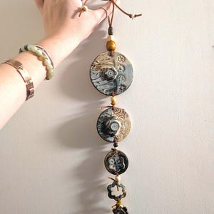 May include: A vertical wall hanging featuring five circular and flower-shaped ceramic pieces in shades of blue, brown, and cream. The pieces are connected by a brown leather cord with wooden and ceramic beads, finished with a bow at the top.