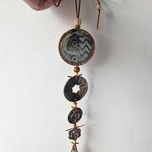 May include: A vertical wall hanging ornament featuring five circular and flower-shaped ceramic pieces in shades of blue, brown, and grey, strung together with brown leather cord and accented with small wooden beads and a top bow.