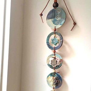May include: A vertical wall hanging featuring four ceramic discs and floral shapes in shades of blue, green, and brown. The pieces are connected by a brown leather cord with wooden beads, finished with a bow at the top for hanging.