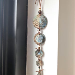 May include: A vertical wall hanging featuring five ceramic pieces in shades of blue and brown, strung together on a brown leather cord. The pieces include circular, flower-shaped, and teardrop forms with etched patterns, separated by small wooden beads.
