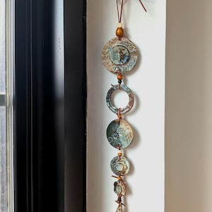 May include: A vertical wall hanging ornament featuring a series of circular and teardrop-shaped ceramic pieces in shades of blue, brown, and cream. The components are connected by brown leather cords and wooden beads, ending in a pointed teardrop shape.