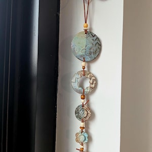 May include: A vertical wall hanging featuring five ceramic discs and flower-shaped pieces in shades of blue, teal, and tan. The pieces are strung together on a brown leather cord with small wooden beads, suspended from a white wall hook.