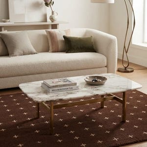 Puede incluir: Una mesa de centro rectangular con una superficie de mármol blanco con vetas grises y una estructura de metal dorado. La mesa está situada en una sala de estar y sostiene dos libros apilados y un cuenco gris poco profundo con objetos metálicos.