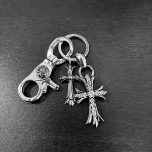 May include: A silver-toned metal keychain featuring a lobster clasp and a split ring. Attached to the ring are two cross pendants: one plain silver cross with intricate detailing and one larger cross encrusted with clear, sparkling stones.
