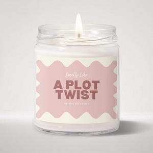 May include: A clear glass jar candle with a lit wick and white wax. The label features a wavy-edged pink rectangle on a cream background with the text Smells Like A PLOT TWIST and NATURAL SOY CANDLE printed in dark brown lettering on the front.