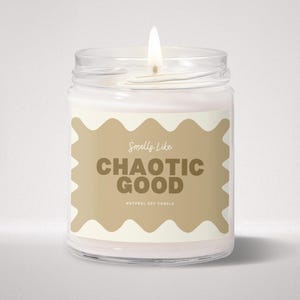 May include: A clear glass jar candle with a lit wick and white wax. The label features a wavy tan border with the text Smells Like CHAOTIC GOOD and NATURAL SOY CANDLE printed in dark brown lettering on the front of the container.