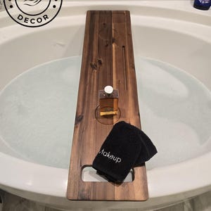 May include: A rectangular dark brown wooden bathtub tray with a smooth finish, featuring a circular cutout for a glass bottle, a long narrow slot, and a rectangular indentation. A black washcloth with the word Makeup embroidered in white sits on top.