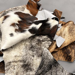 May include: A collection of cowhide leather scraps featuring various natural patterns and textures. The pieces display a mix of brown, black, white, and grey fur, with some sections showing smooth, light-coloured suede undersides visible at the edges.