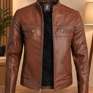 May include: A brown leather jacket featuring a stand-up collar, a full-length front zipper, and two horizontal zippered chest pockets. The interior is lined with black fabric, and a label reading Real Leather is visible at the inner neckline.