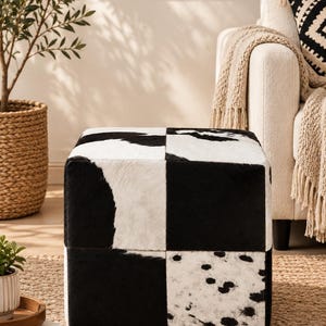 May include: A square ottoman featuring a patchwork design of black and white cowhide panels. The surface displays a mix of solid black sections, white areas with black spots, and irregular black and white patches stitched together in a grid pattern.