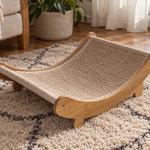 Puede incluir: Una cama para mascotas con un marco de madera curvado en acabado marrón claro y una superficie de descanso de tela beige tejida y texturizada. La estructura se apoya en cuatro patas pequeñas e incluye herrajes metálicos visibles.