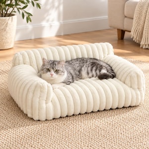 May include: A cream-colored pet bed with a soft, ribbed, corduroy-like texture and raised bolster sides. A grey and white tabby cat with green eyes is lying down on the rectangular cushion, which features a plush, tufted surface design.
