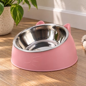 Puede incluir: Un cuenco rosa para mascotas con un inserto de acero inoxidable extraíble. El exterior presenta un diseño de cara de gato moldeado con dos orejas pequeñas en el borde y tres bigotes en el lateral. El cuenco está inclinado y tiene acabado mate.