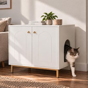 Può includere: Un mobiletto rettangolare bianco per lettiera da gatti con due ante dotate di pannelli incassati ad arco e piccoli pomelli dorati. L'unità poggia su quattro gambe in metallo dorato e presenta un'apertura laterale ad arco per il passaggio.