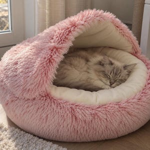 Pode incluir: Uma cama redonda para animais de estimação, cor-de-rosa, em pele sintética, com um topo em forma de capuz e uma almofada interior cor de creme. Um gato de pelo comprido e cor clara com marcas cinzentas dorme enrolado dentro da estrutura.
