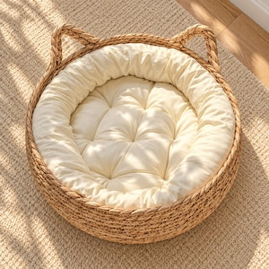 Puede incluir: Una cama redonda para mascotas con una estructura de cesta de fibra natural tejida y dos asas verticales en forma de orejas. El interior contiene un cojín circular extraíble, acolchado y de color blanco roto con un borde suave.