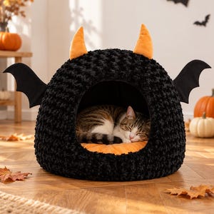 Pode incluir: Uma cama para animais de estimação preta, texturizada e em forma de cúpula, com dois chifres de pelúcia laranja no topo e duas asas de morcego pretas nas laterais. O interior é forrado com uma almofada laranja e um gato malhado dorme dentro.