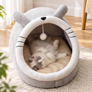Puede incluir: Una cama para mascotas gris en forma de cúpula con dos orejas erguidas en la parte superior y una entrada forrada de felpa blanca con bigotes negros bordados. Un pequeño pompón blanco cuelga del techo interior, que tiene un cojín beige.