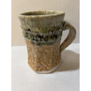 May include: A ceramic mug featuring a textured, tan-colored lower section with small indentations and a smooth, glossy upper rim glazed in shades of sage green and dark brown with a flowing, marbled drip pattern and a sturdy, curved handle.