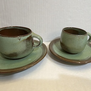 May include: Two ceramic cups and matching saucers featuring a mottled sage green and brown glaze. Each cup has a rounded body with a side handle, and each saucer has a wide, flat rim. The finish shows variations in color and texture throughout.