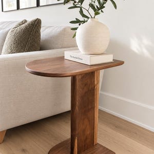 Peut inclure: Une table d'appoint en forme de C fabriquée en bois brun foncé avec un plateau arrondi lisse et une base arrondie assortie reliée par un pilier de support vertical. Un vase en céramique blanche avec des branches d'olivier repose sur un livre.