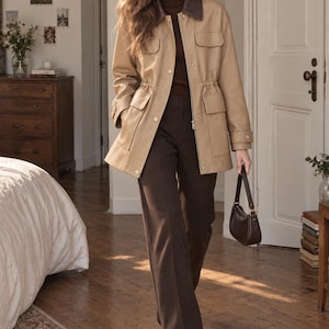 May include: A tan utility jacket with a dark brown collar, featuring a cinched waist, four front flap pockets, and buttoned cuffs. The jacket is worn over a dark brown turtleneck sweater, dark brown trousers, and dark brown boots, with a small handbag.