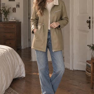 May include: An olive green utility jacket with a cream-colored faux shearling collar, a full-length silver front zipper, a zippered chest pocket, and a drawstring waist, paired with light blue denim jeans and beige ankle boots.