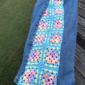 May include: Blue denim jeans featuring a vertical side panel made of light blue crochet granny squares. Each square contains a floral-like pattern in pastel shades of pink, yellow, and lavender, stitched directly into the denim fabric.