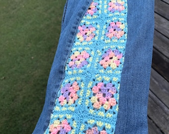 May include: Blue denim jeans featuring a vertical side panel made of light blue crochet granny squares. Each square contains a floral-like pattern in pastel shades of pink, yellow, and lavender, stitched directly into the denim fabric.