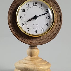 May include: A round analog clock with a dark brown wooden frame and a white face featuring black numerals and hands. The clock is set within a gold-toned bezel and is mounted on a light-colored, tiered wooden pedestal base with a smooth finish.