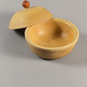 May include: A light-colored wooden bowl with a matching domed lid. The bowl features a visible natural wood grain pattern throughout its surface. The lid is topped with a small, round, dark brown wooden knob handle centered on the top.