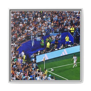 May include: A framed photograph of a soccer player in a white and light blue striped jersey walking toward a corner kick on a green field. A large crowd of spectators in the stands wears matching colors, with blue stadium barriers visible.
