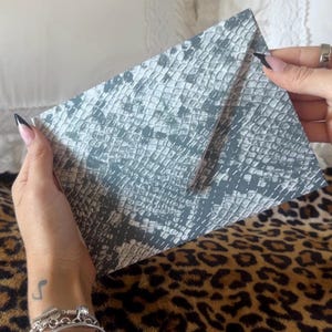 May include: A rectangular envelope featuring a gray and white snakeskin pattern. The envelope has a pointed flap closure and a textured surface that mimics the appearance of reptile scales in a monochromatic color scheme.