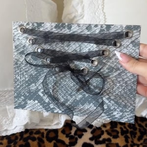 Könnte beinhalten: Ein rechteckiges Stück Material mit grau-weißem Schlangenledermuster und zwei Reihen silberner Metallösen. Ein transparentes schwarzes Band ist kreuzweise durch die Ösen gefädelt und unten in der Mitte zu einer Schleife gebunden.