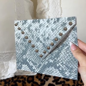May include: A rectangular envelope-style clutch featuring a grey and white snakeskin pattern. The front flap is adorned with a V-shaped row of small, metallic, pyramid-shaped studs along the edge, creating a distinct geometric accent.