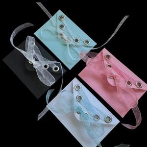 May include: Four rectangular envelopes in black, light blue, pink, and white, each featuring a triangular flap secured with silver metal grommets and tied with a translucent, shimmering organza ribbon threaded through the eyelets.